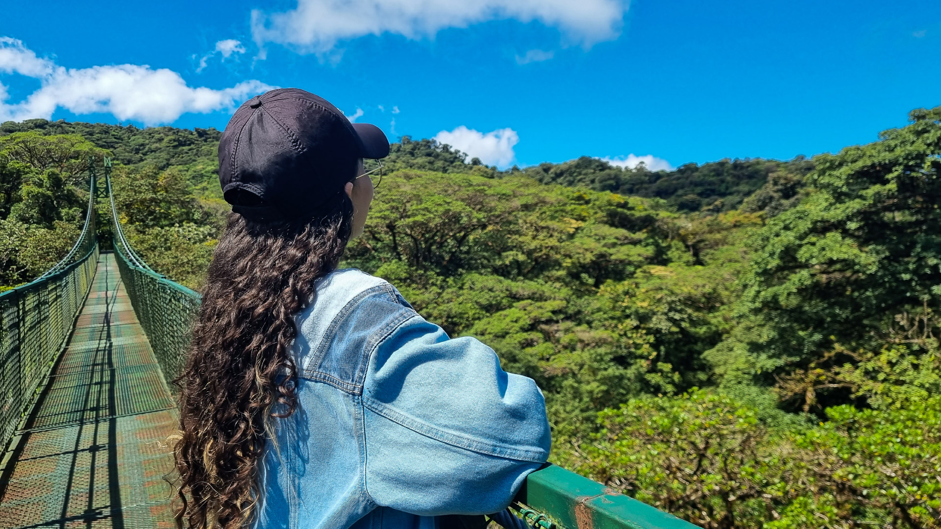 A woman with long curly hair stands on a hanging bridge, enjoying a sunny view of the lush green canopy in Selvatura, Monteverde, Costa Rica.
