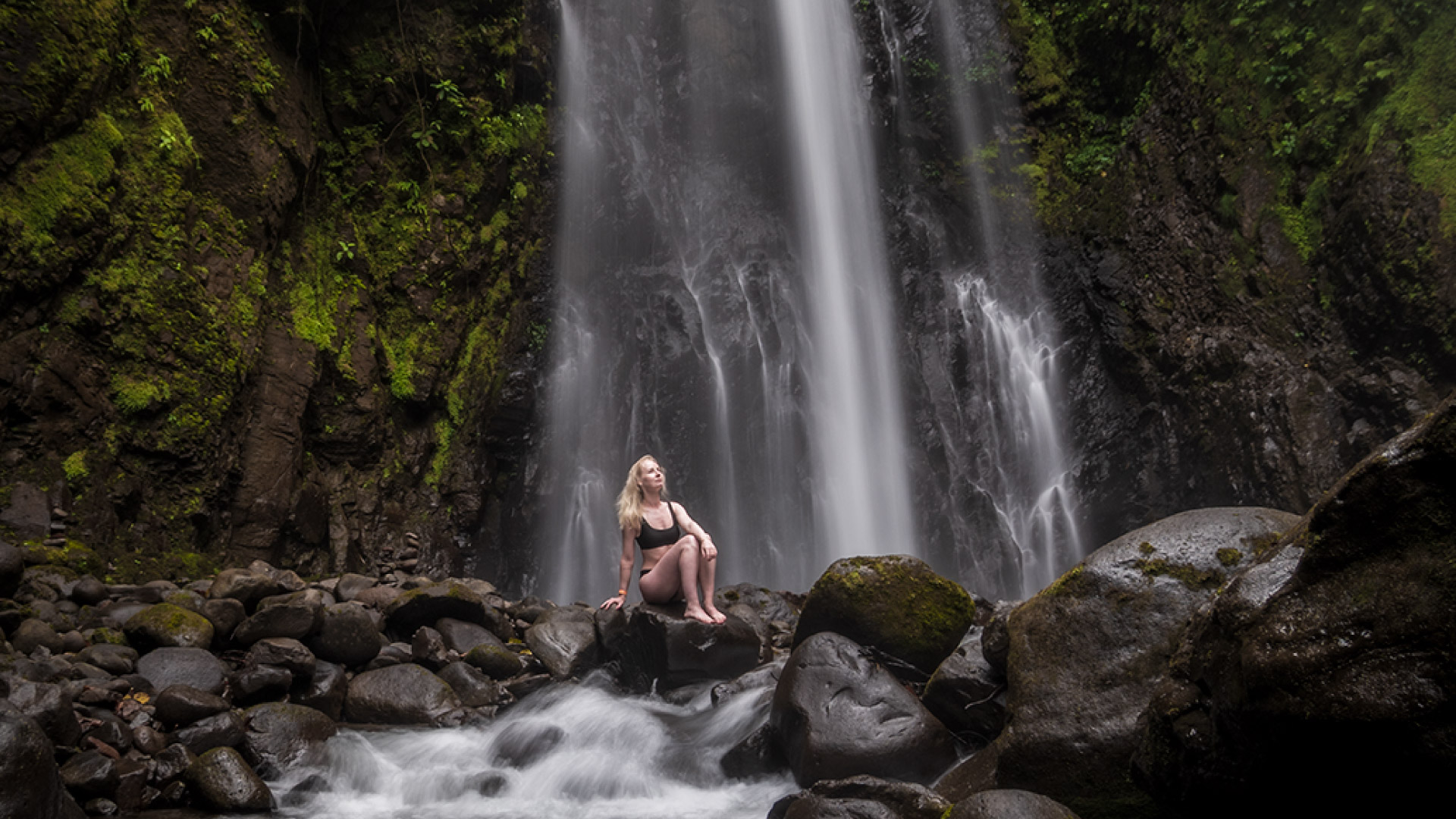 Girl in swimsuit on rocks by river with El Tigre Waterfall in background - Monteverde, Costa Rica